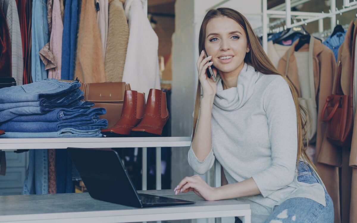 Mulher em boutique fala ao telefone com laptop, cercada por roupas