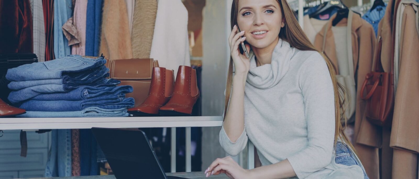 Mulher em boutique fala ao telefone com laptop, cercada por roupas