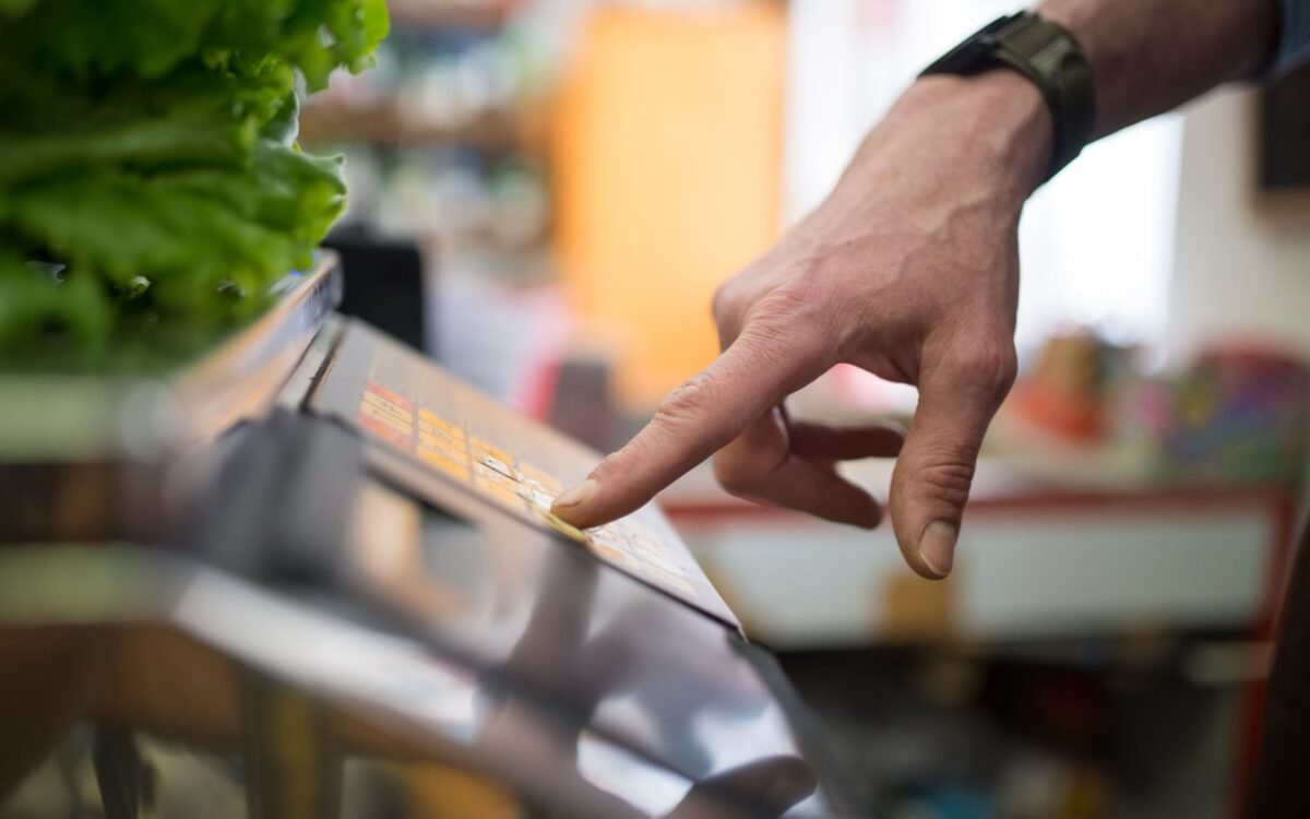 Uma mão interagindo com uma balança digital em uma seção de produtos, enfatizando tecnologia nas compras