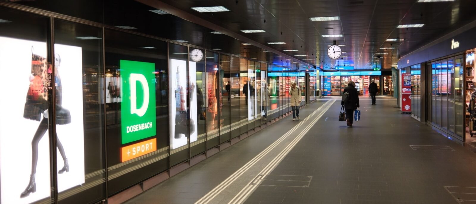 Visão ampla de um corredor de shopping movimentado com pessoas, lojas e um relógio em destaque