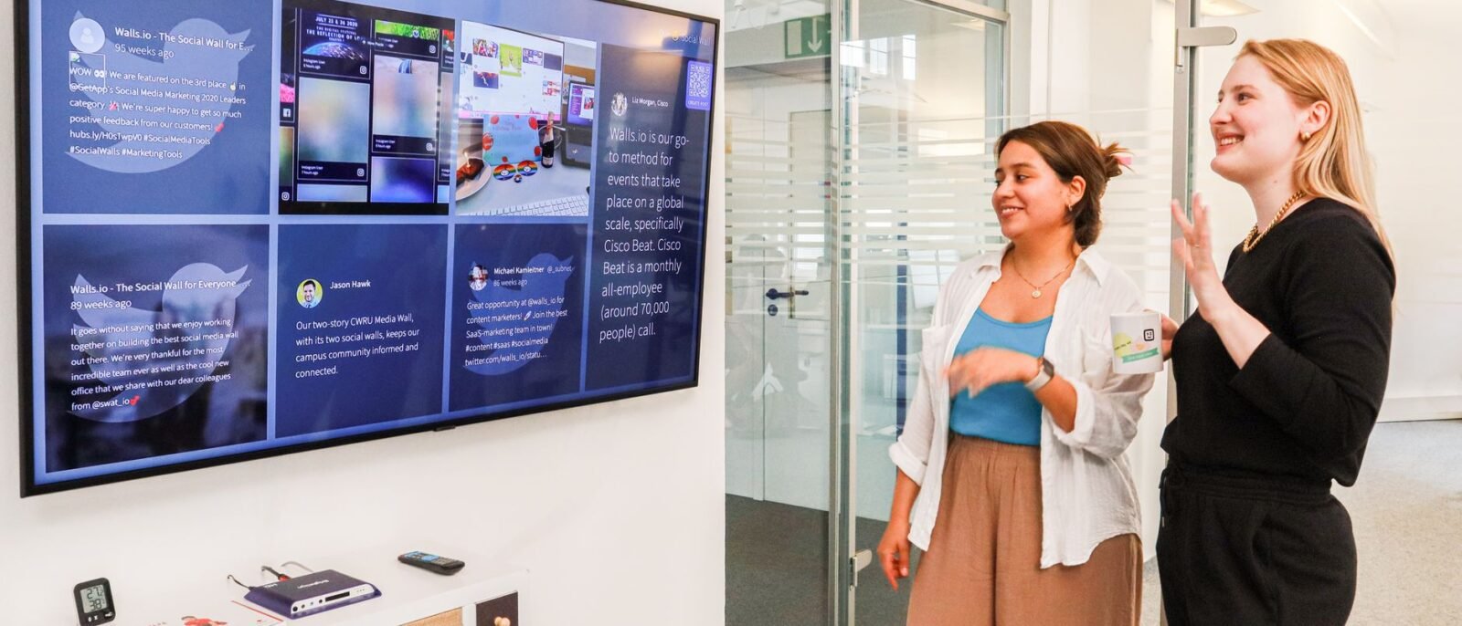 Duas mulheres em um ambiente de escritório interagem com uma grande tela digital exibindo conteúdo de mídia social