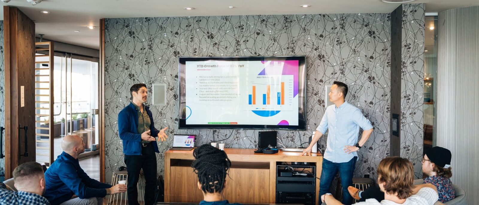 Grupo de profissionais participando de uma apresentação de negócios com uma TV exibindo gráficos